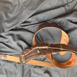 May include: A brown leather belt featuring a wide, reinforced section with multiple brass-toned metal buckles, loops, and studs. The belt has a smooth finish on the exterior and a natural tan leather interior, with a long strap extending from the main body.