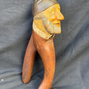 May include: A carved wooden nutcracker shaped like a person's head with a dark brown cap, a light brown face, and a gray beard. The handle features the word BERN carved into the wood. The object has a smooth finish and a distinct facial expression.