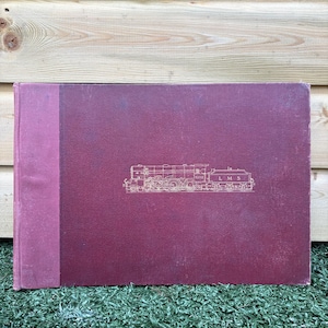 Könnte beinhalten: Ein weinrotes Buch mit einer goldenen Illustration einer Lokomotive auf dem Einband. Das Buch hat einen weinroten Buchrücken und der Text "LMS" ist sichtbar. Das Buch liegt auf grünem Gras.