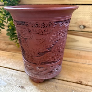 Könnte beinhalten: Ein terrakottafarbener Keramik-Blumentopf mit aufwendigen Drachenschnitzereien. Die detaillierte Reliefarbeit umschließt den zylindrischen Körper des Topfes und zeigt ein traditionelles, asiatisch inspiriertes Design. Der Topf hat einen leicht ausgestellten Rand und einen kleinen Sockel.