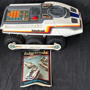 May include: A white and gray Bigtrak programmable toy vehicle with six black wheels, a top-mounted keypad, and a circular hatch. The toy features red, orange, and yellow side stripes and is accompanied by a printed Bigtrak Operator's Manual booklet.
