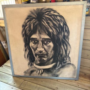 Può includere: Un ritratto quadrato incorniciato che presenta un'illustrazione in bianco e nero in stile carboncino di una persona con capelli lunghi e scalati e una collana con tre ciondoli prominenti a forma di artiglio. La firma dell'artista Nigel è visibile in basso a sinistra.