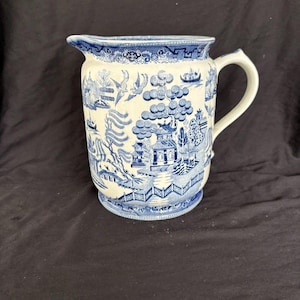 Puede incluir: Jarra de cerámica blanca y azul con asa y pico vertedor. La jarra presenta un diseño detallado de patrón Willow con pagodas, árboles y pájaros. El borde y la base tienen un borde decorativo. La jarra es probablemente vintage o antigua.