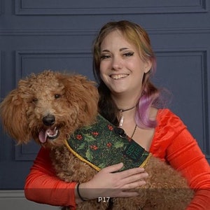 May include: A woman with long, colourful hair is holding a brown and white poodle wearing a green and red festive vest with the text "See Ya Later" on it.