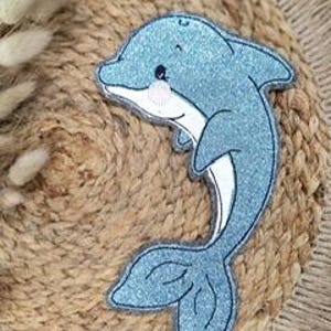 Könnte beinhalten: Ein blauer Glitzer-Delfin-Applikator mit weißem Umriss. Der Delfin blickt nach rechts und hat ein lächelndes Gesicht.