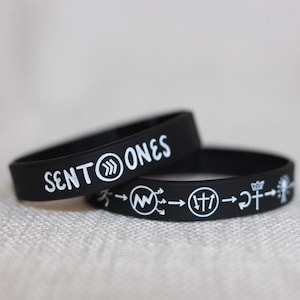 May include: Two black silicone bracelets. The top bracelet has the text "SENT→ONES" in white. The bottom bracelet has a series of white symbols including arrows, a crown, and a cross.