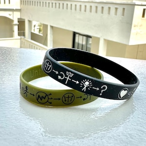 May include: Two silicone wristbands with black and green bands. The black band has white symbols and the text "Until" and the green band has white symbols and the text "27".