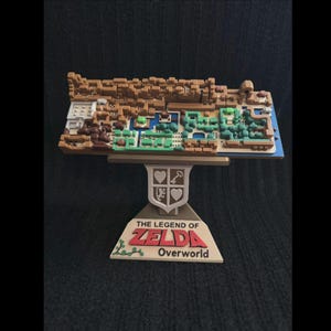 May include: A 3D printed model of the overworld map from The Legend of Zelda for the Nintendo Entertainment System. The map is brown and green and features a variety of terrain, including mountains, forests, and rivers. The map is mounted on a wooden stand with the words "The Legend of Zelda" and "Overworld" printed on it.