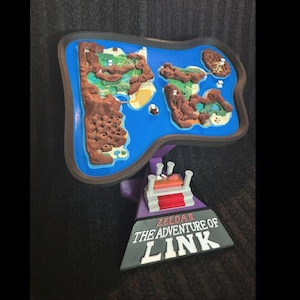 May include: A 3D-printed map of a video game world, featuring a blue sea, brown landmasses, and a decorative base with the text "ZELDA II THE ADVENTURE OF LINK". The map is detailed with various terrain features.