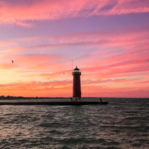 May include: A red lighthouse stands tall against a vibrant pink and orange sunset sky. The lighthouse is situated on a pier extending into a calm body of water.