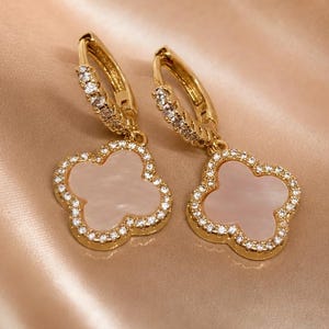 May include: A pair of gold-coloured clover-shaped earrings. Each earring features a four-leaf clover pendant with a light pink centre, surrounded by a border of small, clear crystals. The hoop portion of the earrings is also adorned with crystals.