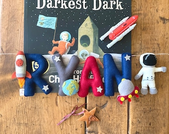 May include: A felt letter banner spelling out the name "RYAN" with a rocket, astronaut, and planets. The banner is hanging above a book titled "Darkest Dark" by Chris and Bert.