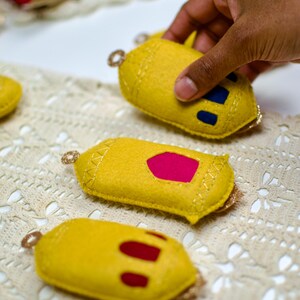Pu&ograve; includere: Ornamenti in feltro giallo con finiture dorate e ritagli colorati. Uno &egrave; tenuto in mano. Gli ornamenti hanno un anello per appenderli. Lo sfondo &egrave; un tessuto color crema a trama aperta.