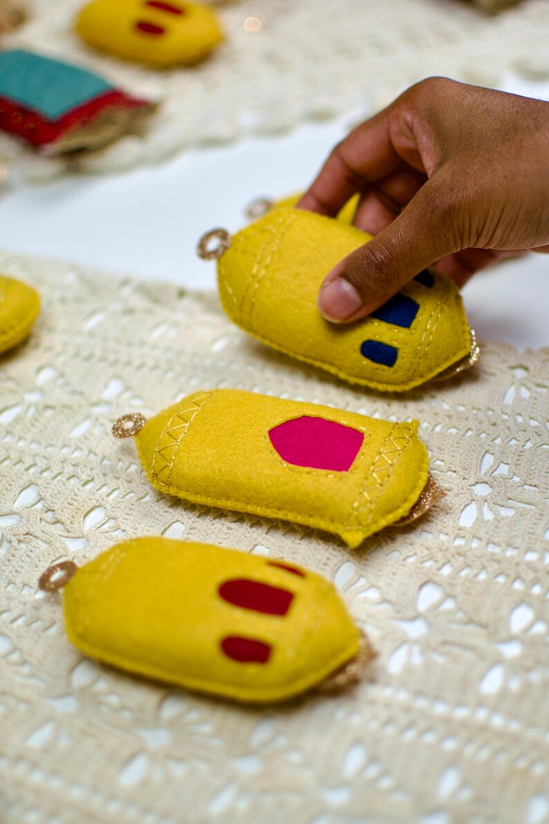 Pu&ograve; includere: Ornamenti in feltro giallo con finiture dorate e ritagli colorati. Uno &egrave; tenuto in mano. Gli ornamenti hanno un anello per appenderli. Lo sfondo &egrave; un tessuto color crema a trama aperta.