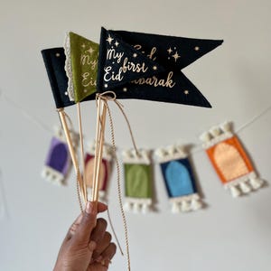 Pu&ograve; includere: Un set di tre bandiere di feltro con lettere dorate. Le bandiere sono nere, verdi e dorate. La bandiera nera dice "Il mio primo Eid Mubarak" con stelle. La bandiera verde dice "Il mio Eid".