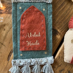 Op de afbeelding: Een gebedsmat van vilt met een rode boog en gouden belettering die "Abdul Hadi" luidt op een teal achtergrond met gouden sterren. De mat heeft een witte franje rond de randen.