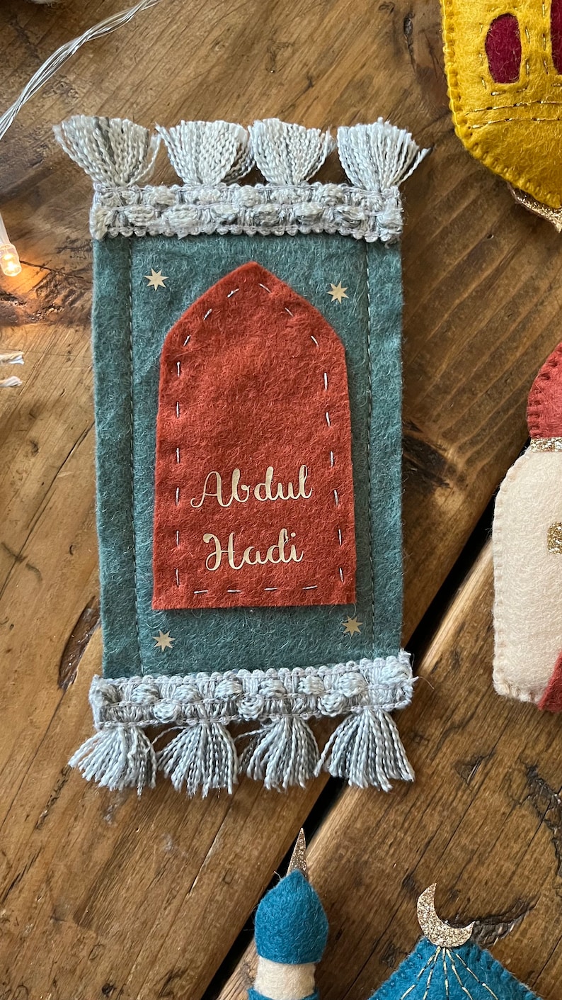 Op de afbeelding: Een gebedsmat van vilt met een rode boog en gouden belettering die "Abdul Hadi" luidt op een teal achtergrond met gouden sterren. De mat heeft een witte franje rond de randen.