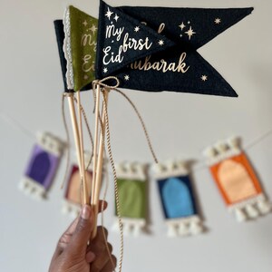 Pu&ograve; includere: Due bandiere di feltro con stelle dorate scintillanti e il testo "My first Eid Mubarak" su una bandiera nera e "My Eid" su una bandiera verde. Le bandiere sono attaccate a bastoncini di legno e tenute in una mano.