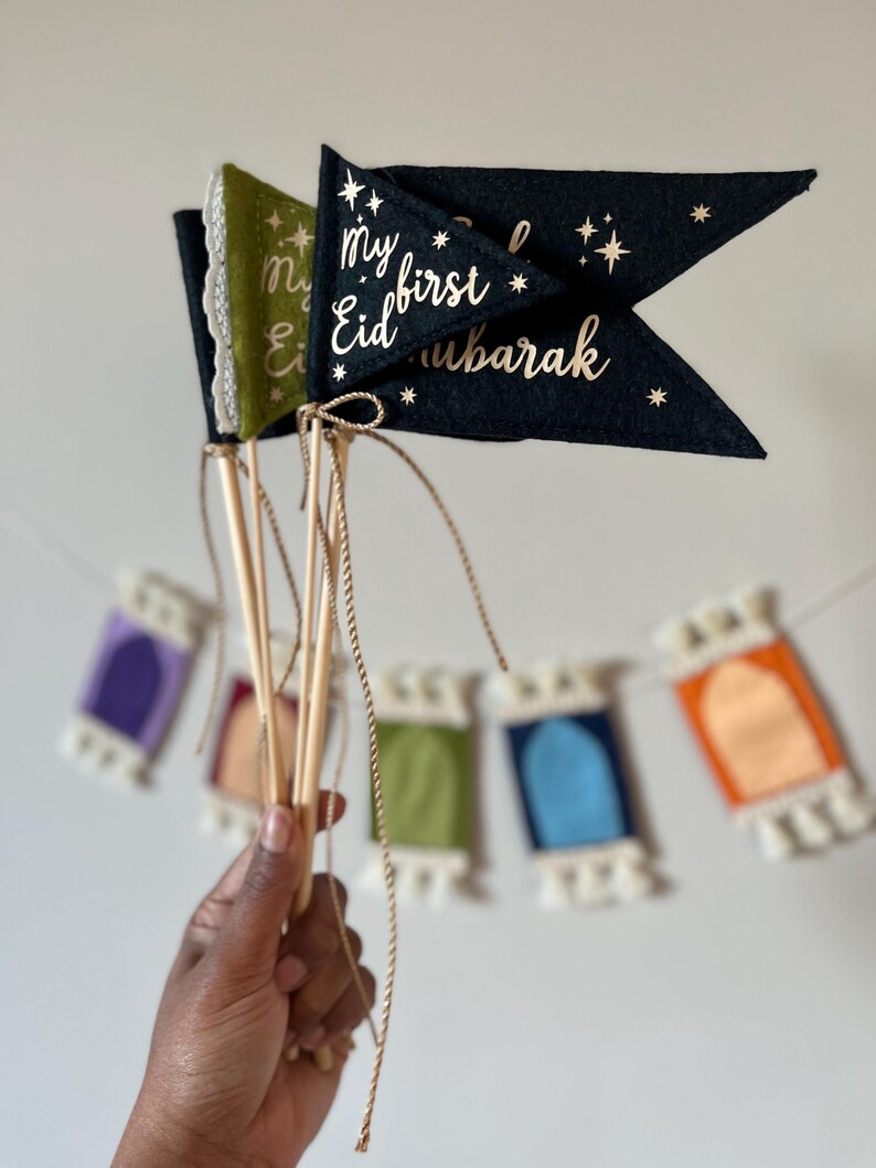Pu&ograve; includere: Due bandiere di feltro con stelle dorate scintillanti e il testo "My first Eid Mubarak" su una bandiera nera e "My Eid" su una bandiera verde. Le bandiere sono attaccate a bastoncini di legno e tenute in una mano.