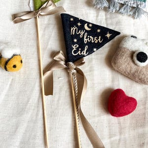 Pu&ograve; includere: Due stendardi in feltro con lettere dorate su un bastone con un nastro marrone. Gli stendardi dicono "my first Eid". Uno stendardo &egrave; verde e l'altro &egrave; nero. Sullo sfondo ci sono un'ape, un cuore e una piccola borsa di feltro.