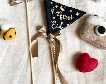 May include: Two felt pennants with gold lettering on a stick with a brown ribbon. The pennants say "my first Eid". One pennant is green and the other is black. There is a bee, a heart, and a small felt bag in the background.