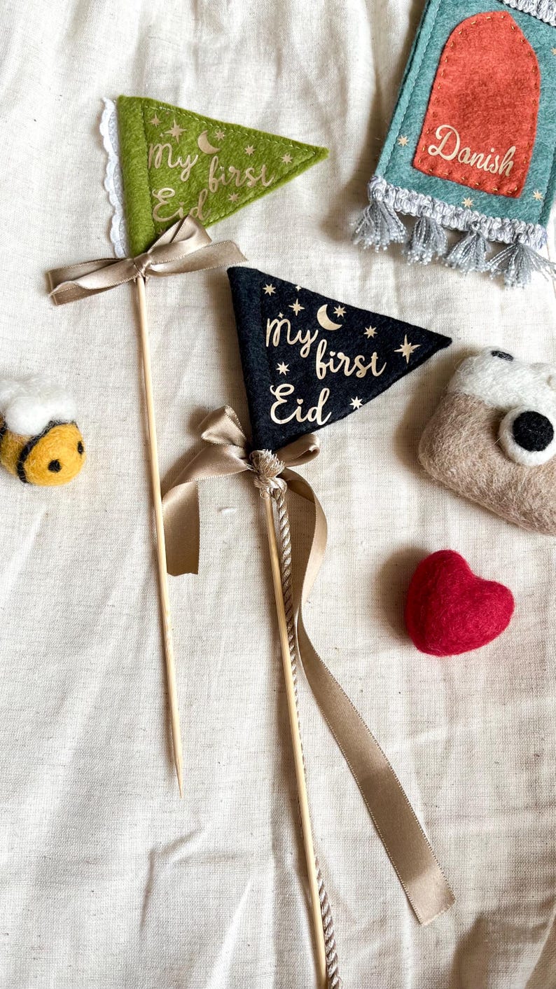 Pu&ograve; includere: Due stendardi in feltro con lettere dorate su un bastone con un nastro marrone. Gli stendardi dicono "my first Eid". Uno stendardo &egrave; verde e l'altro &egrave; nero. Sullo sfondo ci sono un'ape, un cuore e una piccola borsa di feltro.