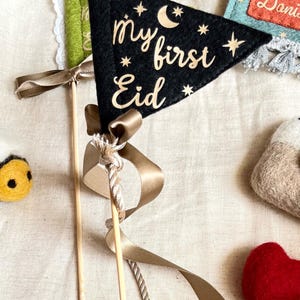 Pu&ograve; includere: Pennant in feltro nero con stelle dorate scintillanti e una luna crescente, con il testo "my first Eid" in lettere dorate scintillanti. Il pennant &egrave; attaccato a un bastone di legno con un nastro marrone.