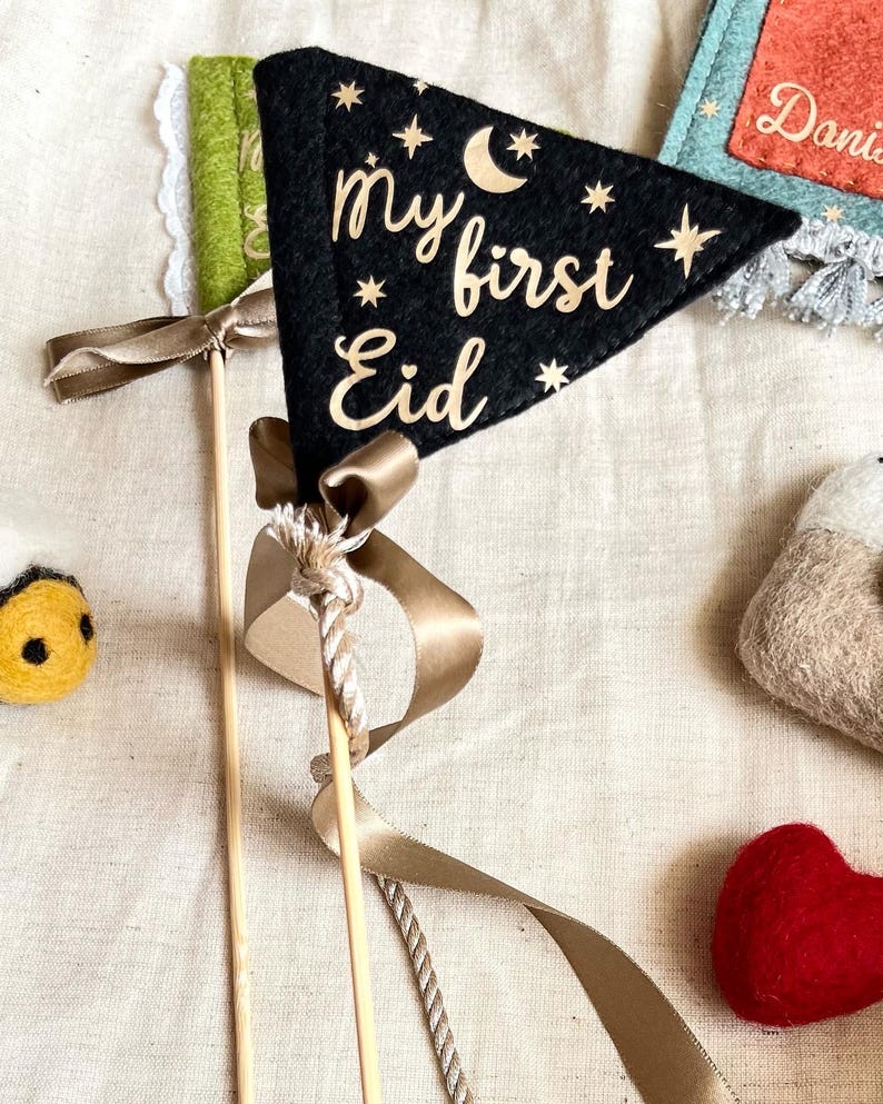 Pu&ograve; includere: Pennant in feltro nero con stelle dorate scintillanti e una luna crescente, con il testo "my first Eid" in lettere dorate scintillanti. Il pennant &egrave; attaccato a un bastone di legno con un nastro marrone.