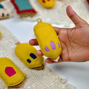 Pu&ograve; includere: Una lanterna fatta a mano in feltro giallo con accenti viola e oro &egrave; tenuta in mano. Altre decorazioni in feltro sono sullo sfondo. La lanterna ha una parte superiore dorata decorativa e un fondo dorato scintillante.
