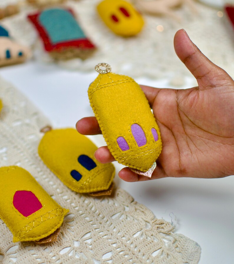 Pu&ograve; includere: Una lanterna fatta a mano in feltro giallo con accenti viola e oro &egrave; tenuta in mano. Altre decorazioni in feltro sono sullo sfondo. La lanterna ha una parte superiore dorata decorativa e un fondo dorato scintillante.