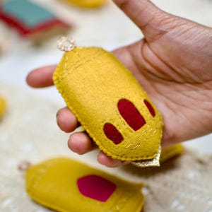 Pu&ograve; includere: Un ornamento fatto a mano in feltro giallo con accenti rossi e finiture dorate. L'ornamento ha un anello per appenderlo ed &egrave; tenuto in mano. Altri ornamenti simili sono sullo sfondo.