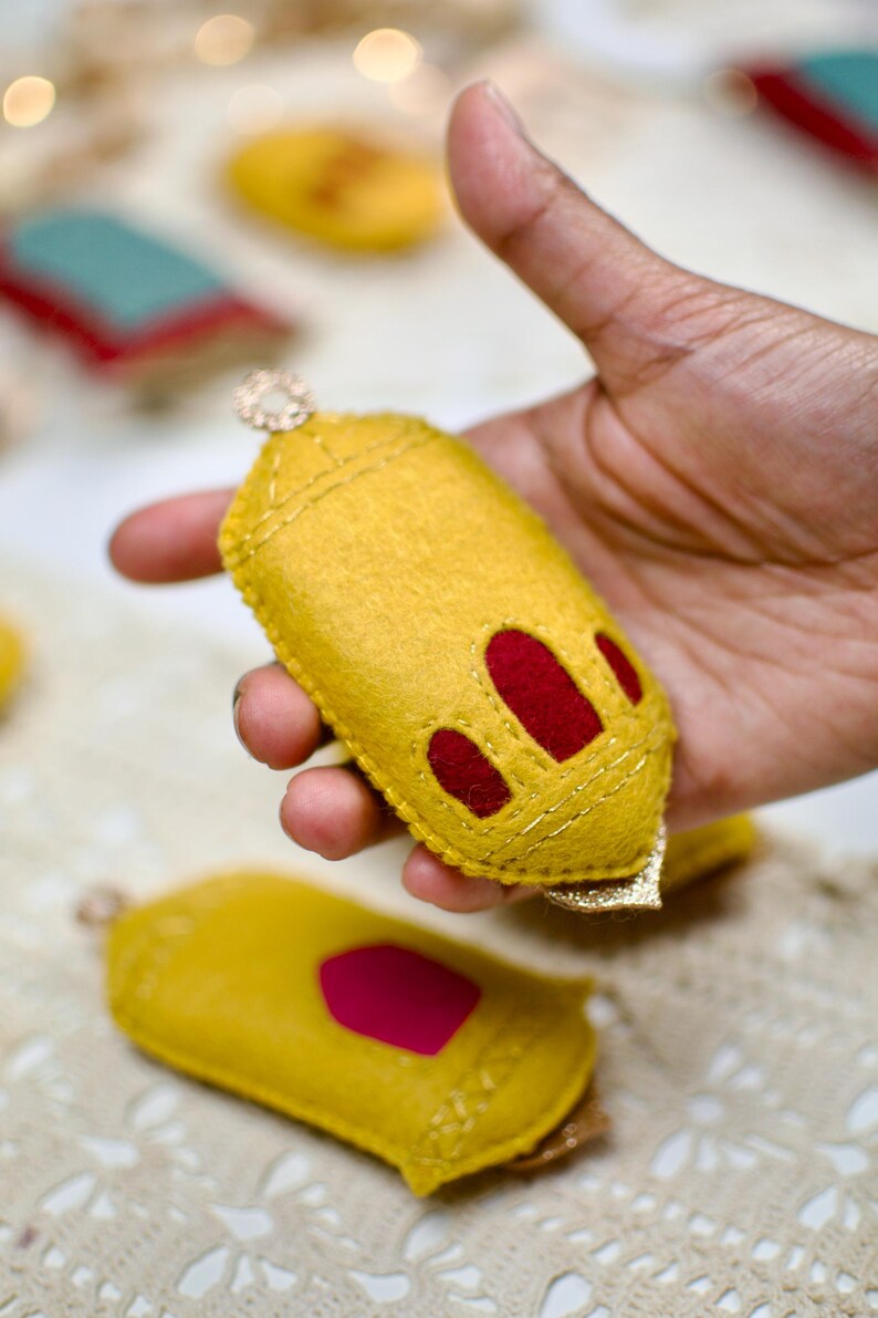 Pu&ograve; includere: Un ornamento fatto a mano in feltro giallo con accenti rossi e finiture dorate. L'ornamento ha un anello per appenderlo ed &egrave; tenuto in mano. Altri ornamenti simili sono sullo sfondo.