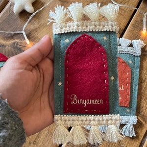 Op de afbeelding: Een viltwerk dat een moskee voorstelt met een rode deur en de tekst "Bunyameen" erop geborduurd. De moskee is versierd met witte kwastjes en sterren.