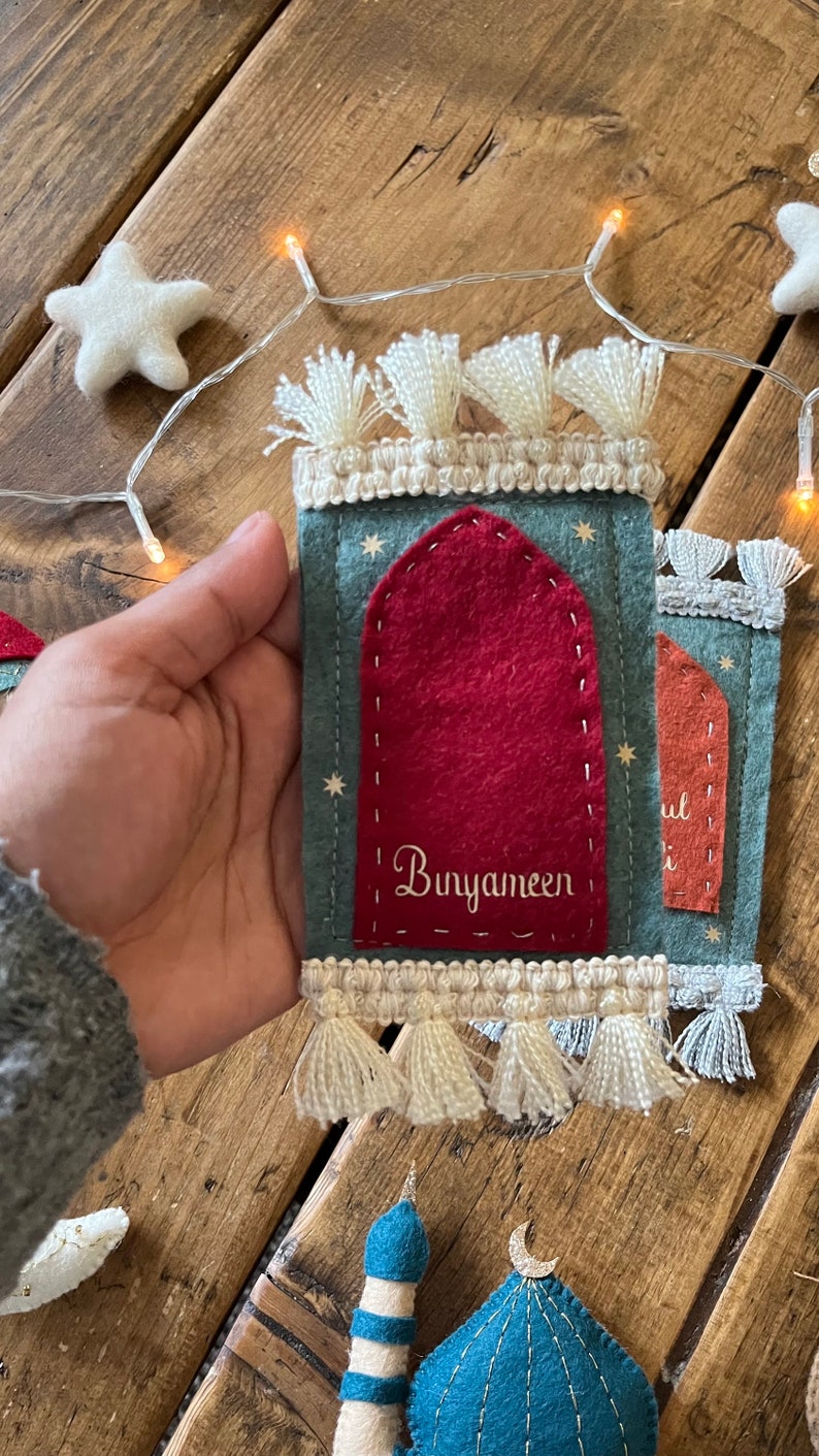 Op de afbeelding: Een viltwerk dat een moskee voorstelt met een rode deur en de tekst "Bunyameen" erop geborduurd. De moskee is versierd met witte kwastjes en sterren.