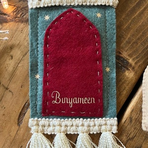 Op de afbeelding: Een groene vilten ornament met een rode vilten boog en witte kwastjes. De ornament heeft gouden sterren en de tekst "Bunyameen" geborduurd.