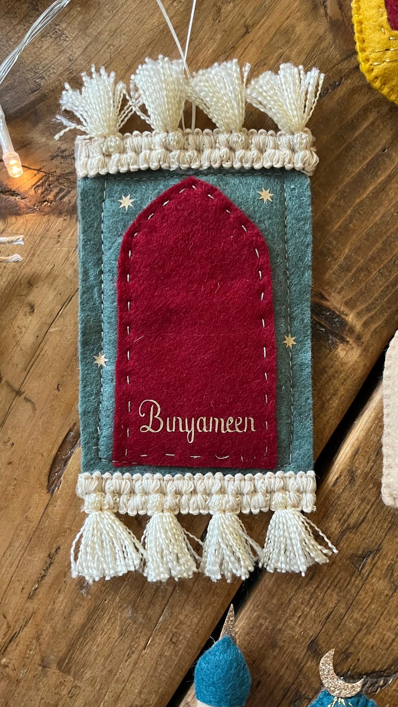 Op de afbeelding: Een groene vilten ornament met een rode vilten boog en witte kwastjes. De ornament heeft gouden sterren en de tekst "Bunyameen" geborduurd.