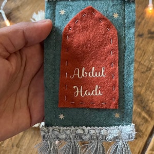 Op de afbeelding: Een tealkleurig gebedskleed van vilt met een rood viltzakje en de naam "Abdul Hadi" geborduurd in wit. Het kleed heeft een grijze kwastrand en is versierd met zilveren glittersterren.