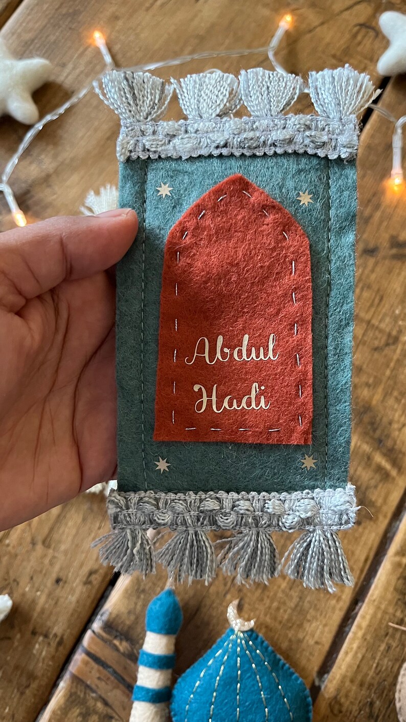 Op de afbeelding: Een tealkleurig gebedskleed van vilt met een rood viltzakje en de naam "Abdul Hadi" geborduurd in wit. Het kleed heeft een grijze kwastrand en is versierd met zilveren glittersterren.