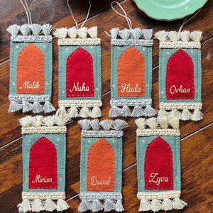 Op de afbeelding: Zeven vilten ornamenten in de vorm van gebedskleden met kwastjes. Elk ornament heeft een andere kleur en een geborduurde naam: Malik, Nuha, Huda, Orhan, Miriam, Dawud en Zyva.