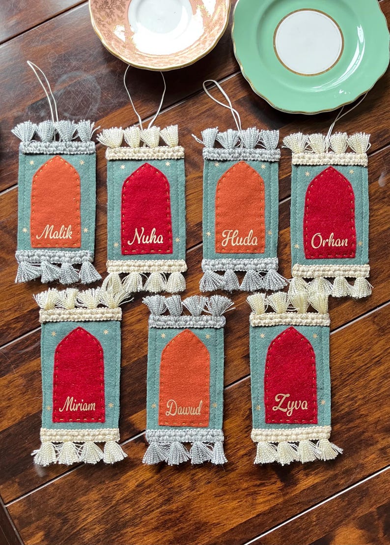 Op de afbeelding: Zeven vilten ornamenten in de vorm van gebedskleden met kwastjes. Elk ornament heeft een andere kleur en een geborduurde naam: Malik, Nuha, Huda, Orhan, Miriam, Dawud en Zyva.