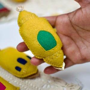 Pu&ograve; includere: Ornamento in feltro giallo con un ovale verde e finiture dorate, tenuto in mano. L'ornamento ha un piccolo gioiello decorativo in cima. Altri ornamenti simili sono visibili sullo sfondo.