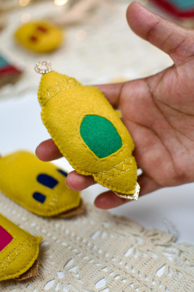 Pu&ograve; includere: Ornamento in feltro giallo con un ovale verde e finiture dorate, tenuto in mano. L'ornamento ha un piccolo gioiello decorativo in cima. Altri ornamenti simili sono visibili sullo sfondo.