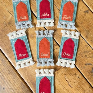 Op de afbeelding: Zeven turquoise vilten ornamenten in de vorm van gebedskleden met witte en grijze kwastjes. Elk ornament heeft een rood of oranje vilten middenstuk met een naam geborduurd in wit: Malik, Nuha, Huda, Miriam, Dawud, Orhan en Zyva.