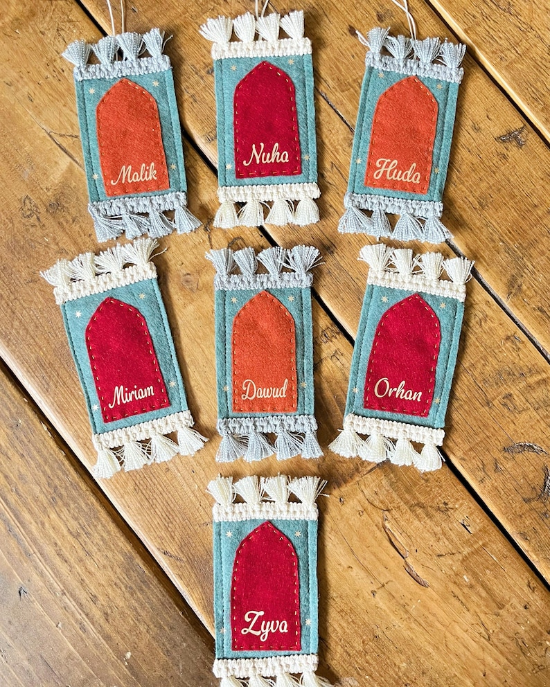 Op de afbeelding: Zeven turquoise vilten ornamenten in de vorm van gebedskleden met witte en grijze kwastjes. Elk ornament heeft een rood of oranje vilten middenstuk met een naam geborduurd in wit: Malik, Nuha, Huda, Miriam, Dawud, Orhan en Zyva.