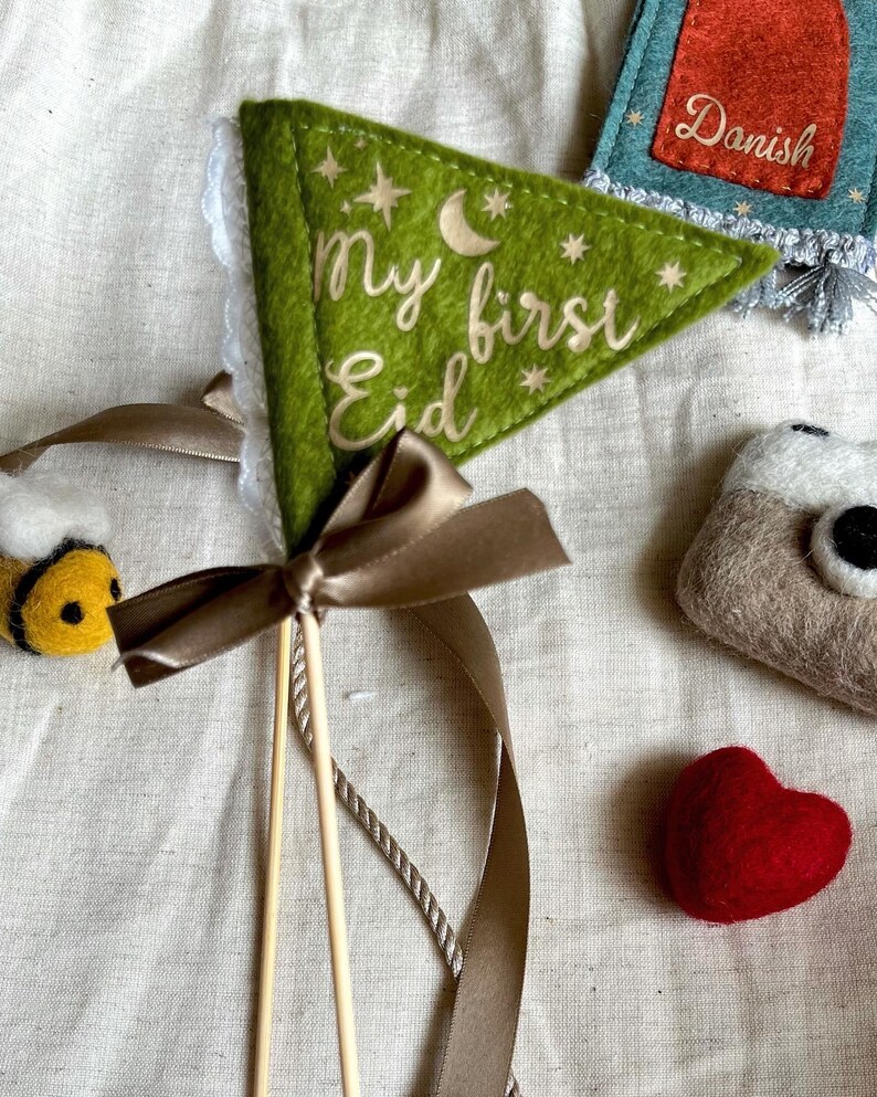 Pu&ograve; includere: Una bandiera triangolare in feltro verde con un bordo in pizzo bianco e un nastro marrone. La bandiera dice "my first Eid" con una luna crescente e stelle.