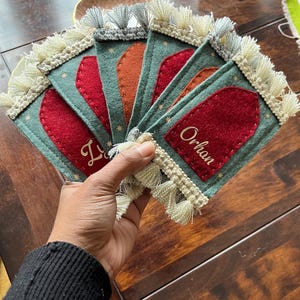 Op de afbeelding: Vijf groene vilt placecardhouders met witte franjes. Elke kaart houder heeft een rode viltinzet met een naam in gouden letters. De namen zijn "Lily", "Orhan", "Lily", "Orhan" en "Lily".