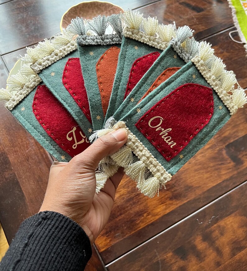 Op de afbeelding: Vijf groene vilt placecardhouders met witte franjes. Elke kaart houder heeft een rode viltinzet met een naam in gouden letters. De namen zijn "Lily", "Orhan", "Lily", "Orhan" en "Lily".