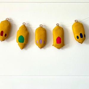 Pu&ograve; includere: Ghirlanda di cinque ornamenti a forma di lanterna in feltro giallo. Ogni lanterna ha una finestra colorata diversa, tra cui rosso, verde, viola e blu. Le lanterne sono sospese a una corda bianca e hanno un piccolo anello dorato nella parte superiore.