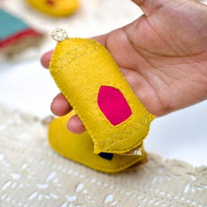 Pu&ograve; includere: Un ornamento fatto a mano in feltro giallo con un inserto rosa e cuciture dorate, tenuto in mano. L'ornamento &egrave; sormontato da un piccolo dettaglio scintillante. Il testo "Swipe to see more designs" &egrave; visibile in basso.