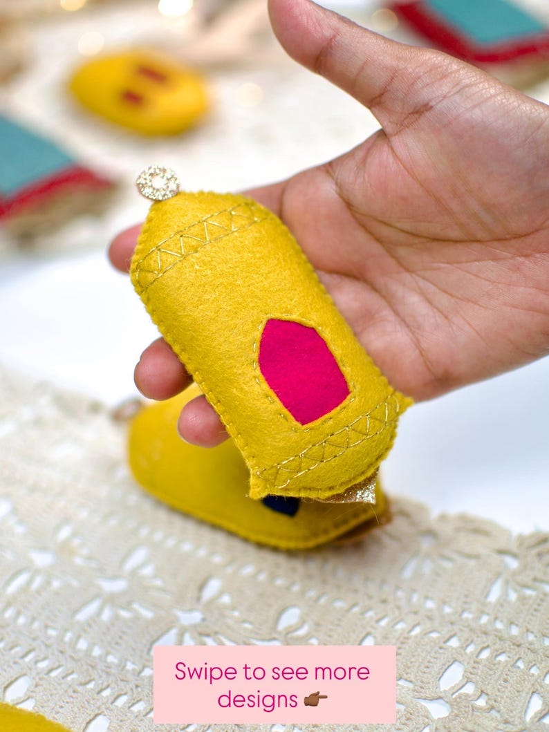 Pu&ograve; includere: Un ornamento fatto a mano in feltro giallo con un inserto rosa e cuciture dorate, tenuto in mano. L'ornamento &egrave; sormontato da un piccolo dettaglio scintillante. Il testo "Swipe to see more designs" &egrave; visibile in basso.